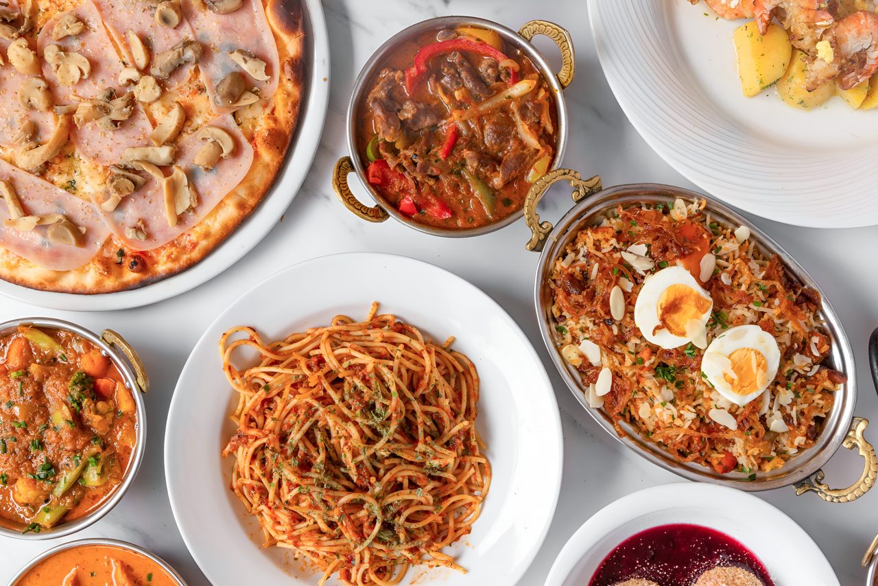 Pizza mit Schinken und Champignons, indische Currys in Kupferschalen, Spaghetti und Hühner-Biryani auf dem Tisch im Café Hufeisen